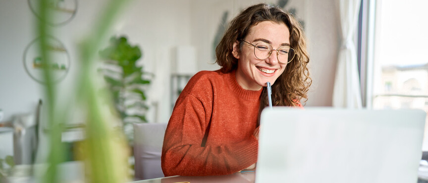 Junge Studentin mit lockigen Haaren, Brille und orangefarbenem Pullover, die vor einem Laptop am Schreibtisch sitzt und an einer Videokonferenz teilnimmt.