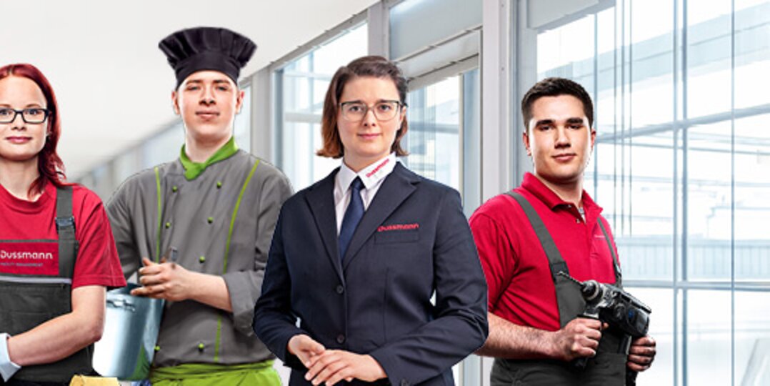 Gruppenbild von vier lächelnden Dussmann-Auszubildenden und Studierenden in Arbeitskleidung aus unterschiedlichen Arbeitsbereichen vor weiß-blauem Hintergrund.