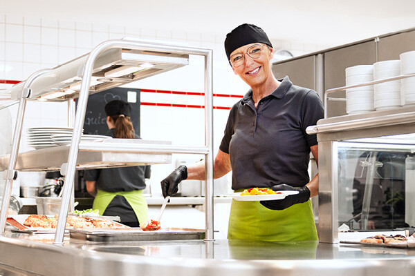 Dussmann-Mitarbeiterin aus der Gastronomie, die gerade in der Kantine Essen ausgibt und lächelt.