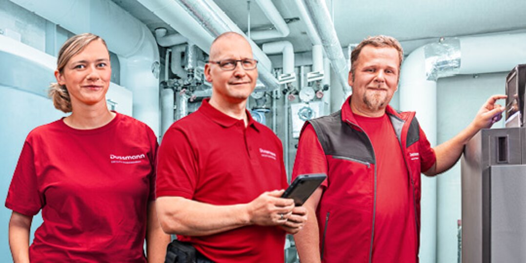 Gruppenbild von drei lächelnden Dussmann-Mitarbeitenden aus dem Arbeitsbereich Gebäudetechnik und Elektrotechnik vor blauen  Hintergrund. 