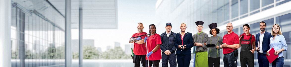 Gruppenbild von lächelnden Dussmann-Mitarbeitenden aus unterschiedlichen Arbeitsbereichen vor neutralem grauem Hintergrund. 