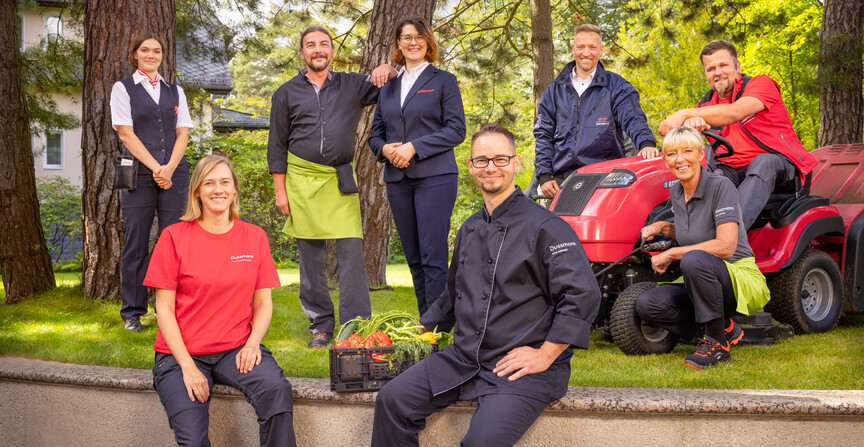 Gruppenbild von lächelnden Dussmann-Mitarbeitenden aus unterschiedlichen Arbeitsbereichen, die in einer Art Vorgarten mit Wiese und Bäumen stehen und ihre Arbeitskleidung tragen. 