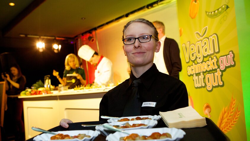 Eine sympathische Frau präsentiert Currywurst auf einem Teller. | © Dussmann Group/Foto: Kay Herschelmann